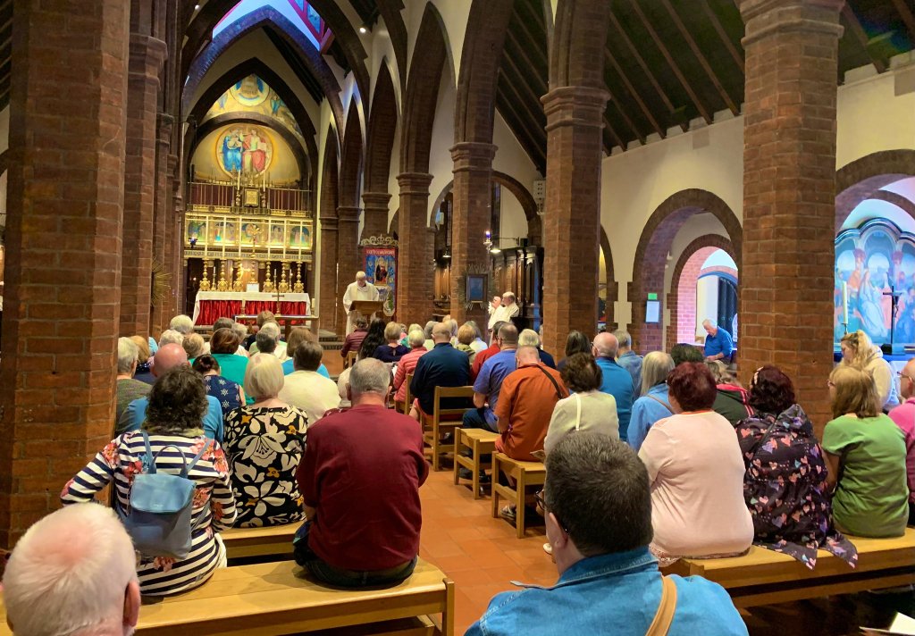 a photo taken from the back of a church which is full of people worshipping and praying. There is a male priest standing at the front of the alter talking. 